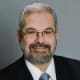 A headshot of a smiling man with short salt and pepper hair and a beard is wearing glasses, a black suit, and a light blue tie.