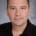 A headshot of Dr. Michael Kruer. A smiling man with short dark brown hair wearing a black collared shirt.