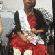 A smiling young woman with cerebral palsy is seated in her wheelchair while wearing a red dress and pageant sash.