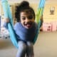 A laughing young girl with CP, wearing blue fleece top, grey leggings, and pink ankle foot orthotics, swings in a blue hammock.