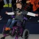 A beaming child with cerebral palsy is seated in a purple wheelchair at a sports venue with arms spread out in celebration.