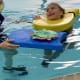 A smiling girl with cerebral palsy swims in a pool with the help of yellow and blue pool floats supported by an instructor.