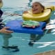 A smiling girl with cerebral palsy swims in a pool with the help of yellow and blue pool floats supported by an instructor.