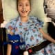 A smiling young girl with CP, wearing a nasal cannula and a hospital gown, reclines in a wheelchair while holding a soft toy.