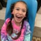 A beaming child with cerebral palsy, wearing a tie-dye shirt, sits in a blue plastic chair.