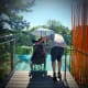 An individual with cerebral palsy is seated in a wheelchair under an umbrella on a sunny deck overlooking trees with an adult.