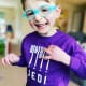 A smiling young boy with cerebral palsy wearing blue glasses and a purple sweater.