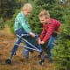 At a tree farm, a young boy with CP sits on his wheeled blue walker while another young boy pulls it from the front.