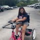 A woman with CP, wearing sunglasses, a navy-blue t-shirt, and grey shorts, rides a pink adaptive tricycle in a parking lot