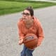 A woman with CP, wearing glasses, an orange hoodie, and jeans, prepares to throw a basketball while outside on her driveway.