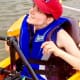 A smiling woman with cerebral palsy, wearing a red baseball cap and royal blue lifejacket, looks up from her adaptive kayak.