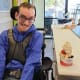 A smiling man with cerebral palsy is seated in his wheelchair in an ice cream parlor next to an ice cream sundae.