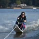 A beaming woman with CP, wearing sunglasses and a black wetsuit, churns up the water as she enjoys adaptive water skiing.