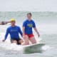 A man, together with a smiling woman with CP, kneel on the same surfboard while gilding through the ocean waves.