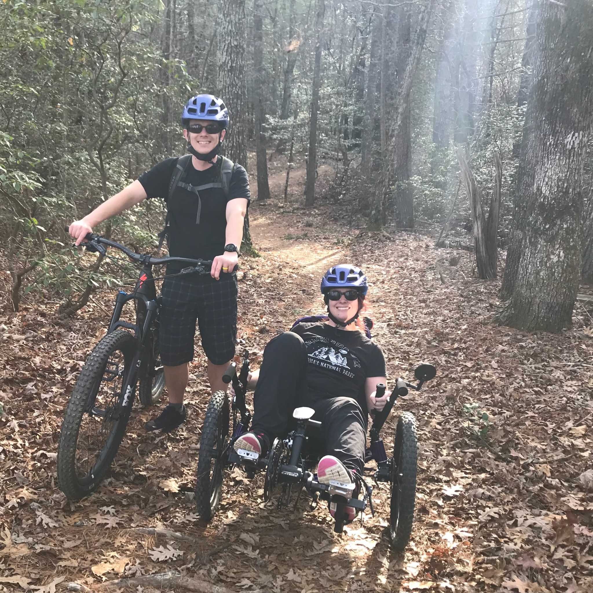 A man standing with a bicycle, and a woman with CP riding a recumbent trike, smile together on a sunlit forest path.