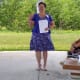 A smiling woman with CP, wearing a blue blouse and dress, holds a sheet of paper while making a speech in an outdoor pavilion.