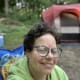 A smiling woman with CP, wearing a pale green sweater and glasses, sits in front of a campsite in a forest clearing.