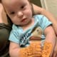 A baby with CP, wearing a blue otter t-shirt and beige sea life kinesiology tape on her hands, lies in her mother’s arms.