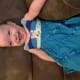 A laughing baby with cerebral palsy, wearing a blue dress, sits on a brown couch.