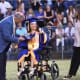 A proud young woman with CP, seated in a wheelchair and wearing a blue-and-gold graduation cap and gown, receives her diploma.