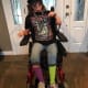 A smiling young woman with cerebral palsy, wearing a decorative eye mask, is seated in her wheelchair by the front door.