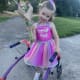 A young girl with CP, wearing a sparkly pink dress, stands outside with her purple walker while holding a bouquet of flowers.