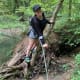 A smiling young woman with cerebral palsy leans against a fallen tree while hiking in a leafy forest next to a shallow stream.