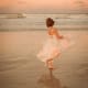 A young woman with CP, wearing a pink-and-white dress, dances on a tide-washed sunset beach facing the rolling ocean waves.