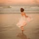 A young woman with CP, wearing a pink-and-white dress, dances on a tide-washed sunset beach facing the rolling ocean waves.