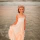 A young girl with cerebral palsy smiles while holding the bottom of her dress above an incoming ocean tide at sunset.