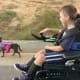 A boy with cerebral palsy using a powerchair smiles down at his small black dog as they walk down a street together.
