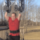 A man with CP, wearing a red t-shirt, support belt, and black sweatpants, lifts his wheelchair above his head on his deck.