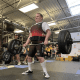 A man with cerebral palsy, wearing a red squat suit, determinedly lifts a large barbell in a gym.