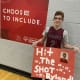 A smiling boy with CP stands in front of a ‘Choose to Include’ banner while holding a sign that reads ‘Hit the shot Ryland’.