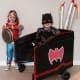 A smiling child with cerebral palsy uses a walker while wearing a Batman costume next to another child also in costume.
