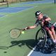 A boy with cerebral palsy leans sideways in his wheelchair to hit a ball with his racket while playing on a tennis court