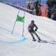 A skier with CP, wearing a grey snowsuit, leans into a sharp turn around a ski gate in an adaptive competition.