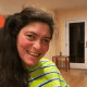 A broadly smiling woman with cerebral palsy, wearing a yellow-striped shirt, in a brightly lit room.