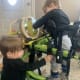 A pair of twins, one a toddler with cerebral palsy in a standing frame, play on a drum set.