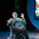 A woman with cerebral palsy is wearing glasses and a sparkling blouse. She is seated in a wheelchair while singing on a stage.