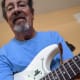 A smiling man with cerebral palsy wears glasses and a blue t-shirt. He is playing a white guitar.