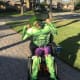 A young man with cerebral palsy is seated in a powerchair in a driveway. He is wearing green face paint and a ‘Hulk’ costume.