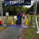 A man with CP, seated in his wheelchair, punches the air in triumph while crossing under the finish line banner of a park race.