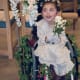 A smiling girl with cerebral palsy, wearing a dress, holds a bouquet while sitting in a wheelchair decorated with ivy leaves.
