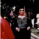 A smiling woman with CP, wearing dark sunglasses and a black graduation cap and gown, proudly holds her college diploma.