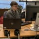 A woman with CP, with short hair, glasses, and a grey blazer, works at her office desk with a laptop and computer monitor.