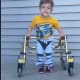 A smiling young boy with cerebral palsy, wearing a yellow t-shirt, stands with his gold walker in front of white home siding.
