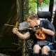 In a forest, a man with cerebral palsy concentrates on firing an arrow from a bow while seated in his powerchair.