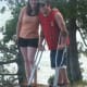 A smiling man with cerebral palsy, using crutches, stands together with a woman on a rocky outcrop overlooking trees and a lake