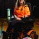 A woman in a bright orange shirt with glasses and long blond e hair smiles in her power wheelchair